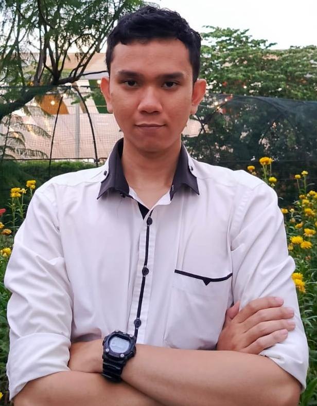 Portrait of Andi Muhammad Naufal with arms crossed, wearing a white shirt with a dark collar and a black digital watch, in front of yellow flowers.